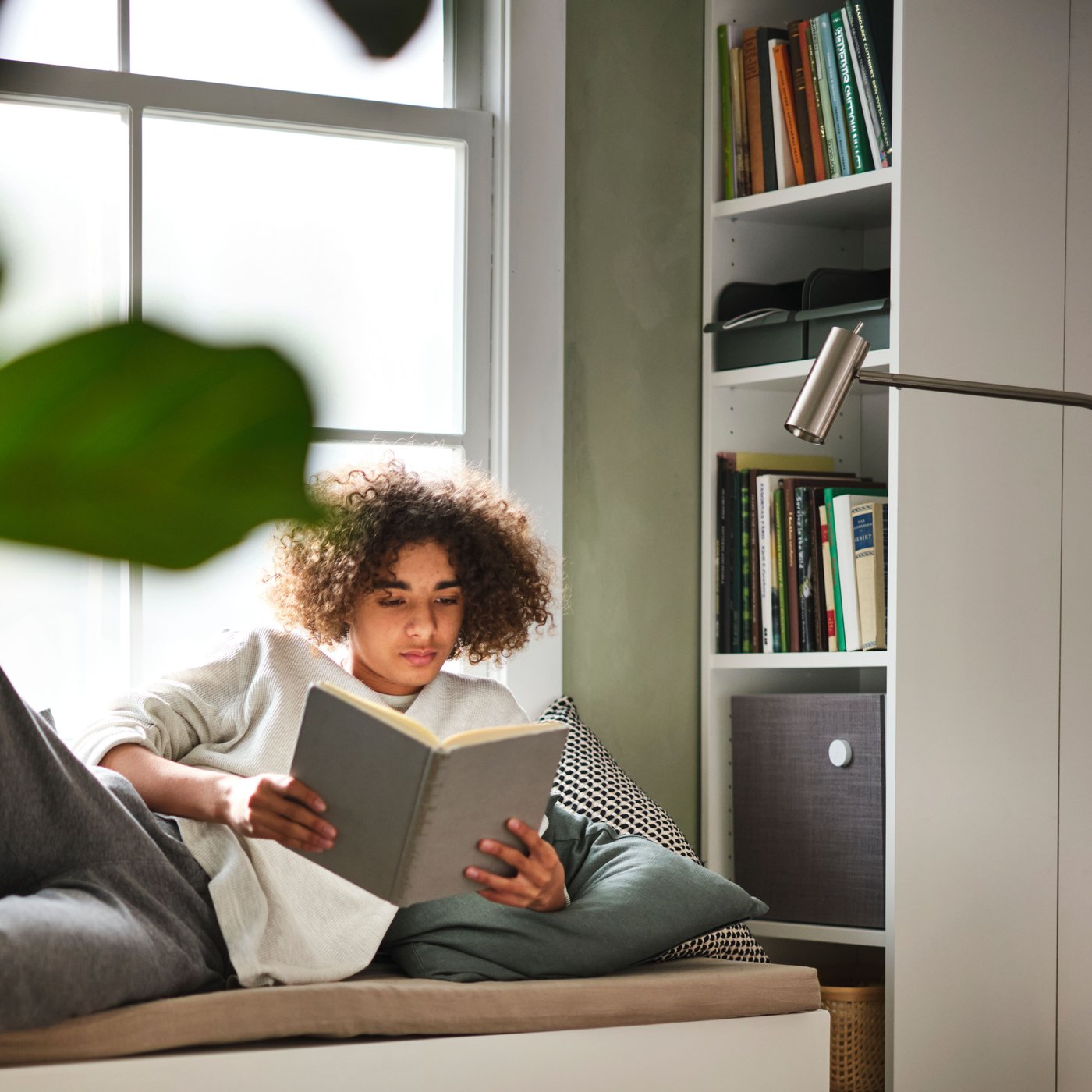 A bright room where a young person reading, lying sideways on a SMÅSTAD bench next to a window and by a bookcase.