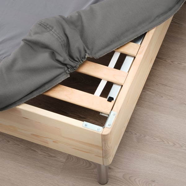 The corner of a LYNGÖR slatted mattress base with legs has the dark grey cover pulled back to show the solid-pine slats.
