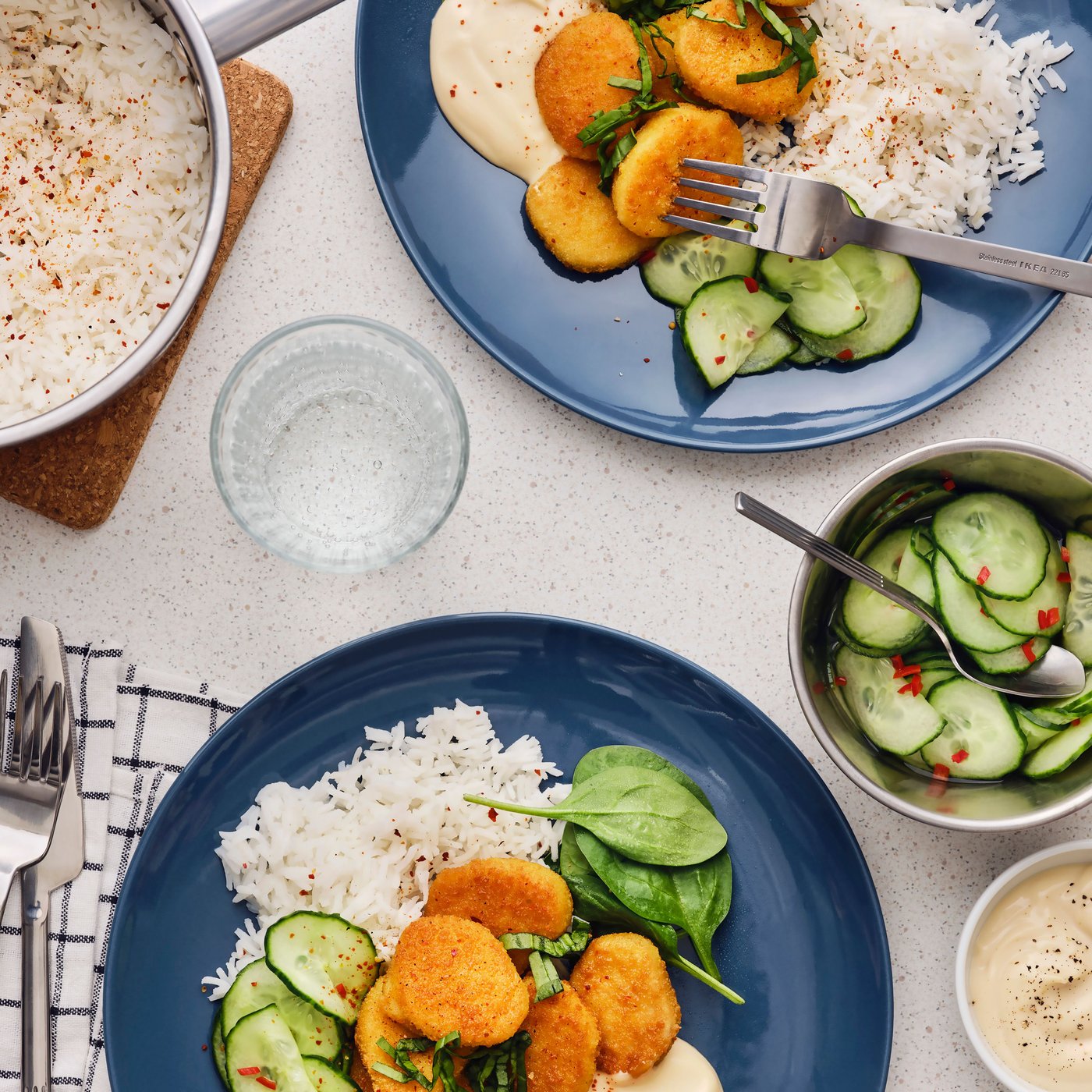 A well-balanced meal setup on a table with plates of rice, cucumber salad, and breaded cutlets on spinach leaves. A glass of water and cutlery complete the scene.