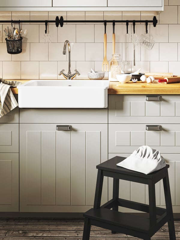 A black step stool with a chef’s hat, beige kitchen fronts, a sink and baking tools on the worktop, black rails.