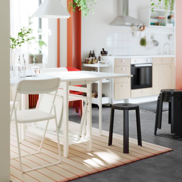 A bright kitchen features a white dining table surrounded by two chairs, with a black stool nearby, and greenery in the background.