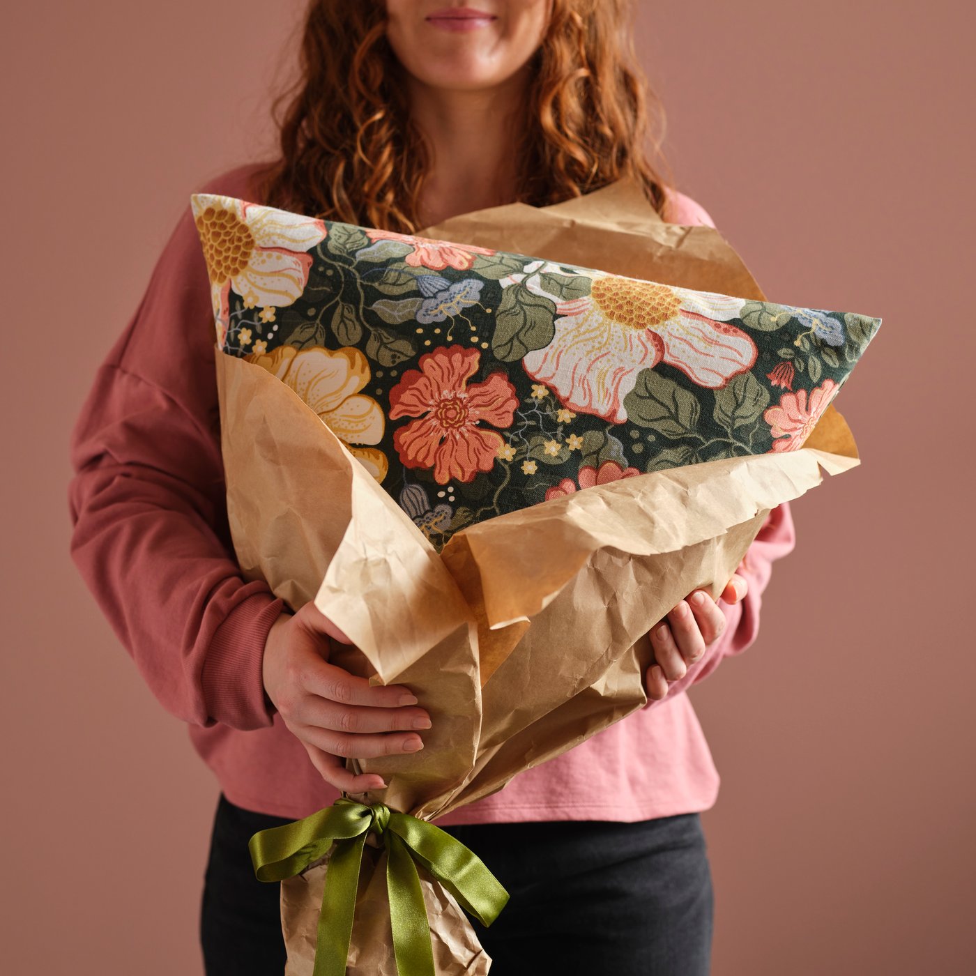 A person in a pink room holds a cushion with a floral ARENBOK cushion cover wrapped in brown paper with a green ribbon.