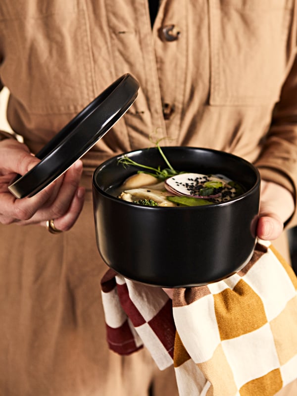 A woman is holding a KRÖSAMOS serving bowl in one hand and small plate in the other. In the bowl is a warming broth.