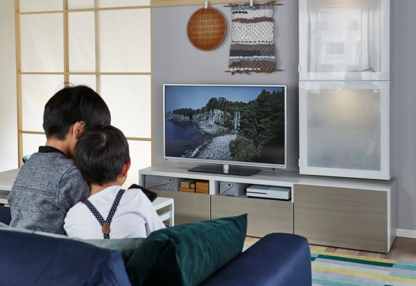 Zwei Kinder sitzen auf einer Couch und sehen fern. Ein Flachbildfernseher steht auf einem TV-Möbel.