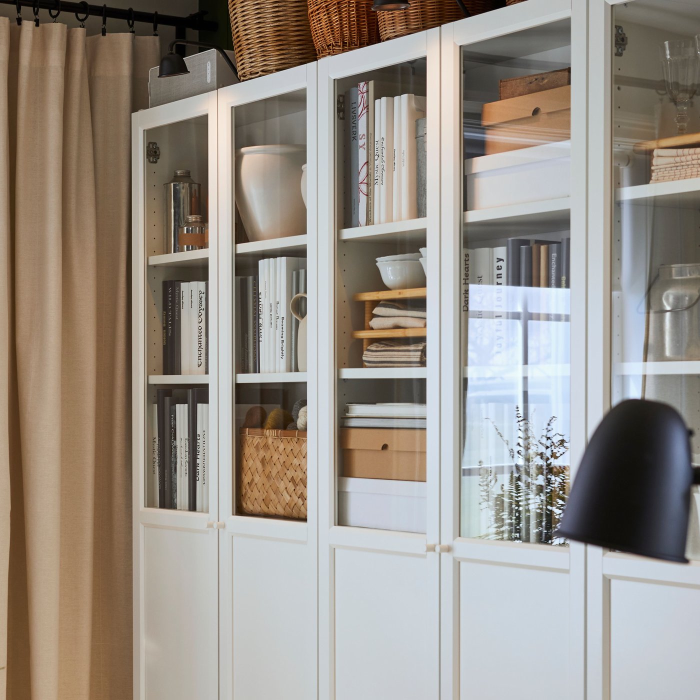 Librerie BILLY con pannelli e ante a vetro, libri e contenitori all'interno e cestini in cima, accanto a una tenda beige.