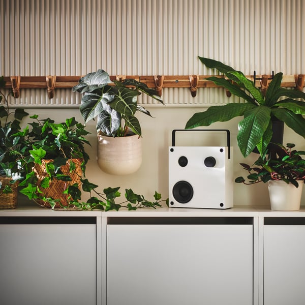 A white VAPPEBY Bluetooth speaker on top of a BISSA shoe cabinet surrounded by various FEJKA artificial potted plants.