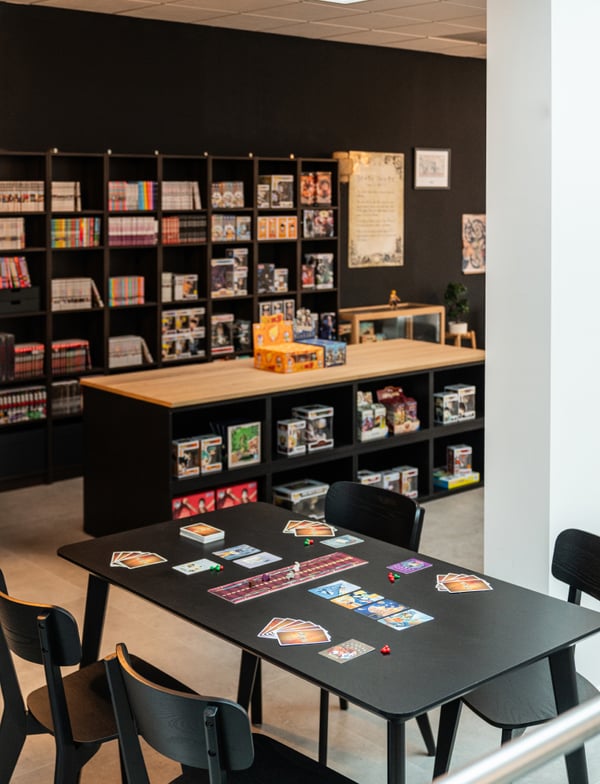 Un tavolo su cui sono disposte carte da gioco, sullo sfondo una libreria e una gondola espositiva della serie BESTÅ.