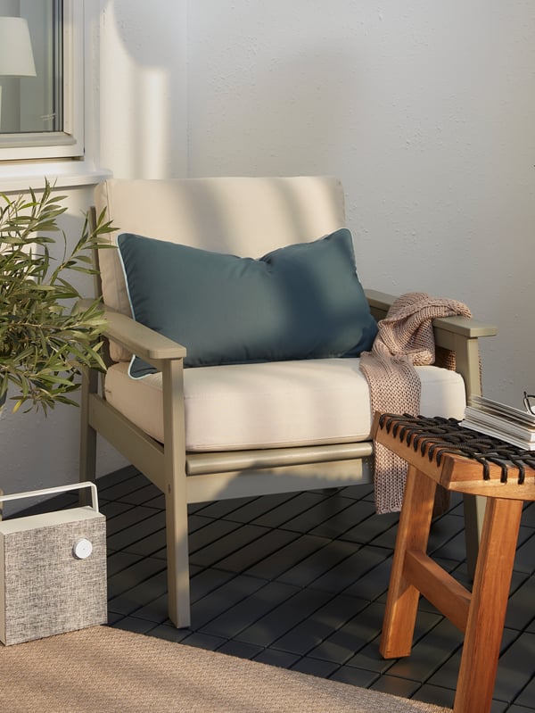 Coin de terrasse accueillant avec fauteuil gris et coussins de chaise, tabouret en bois, enceinte blanc/gris et plaid.