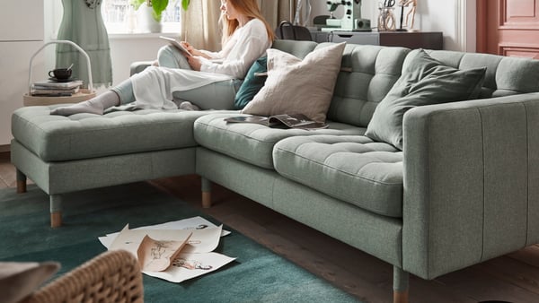 A woman is sitting on a light green LANDSKRONA sofa with a chaise longue in the living room.