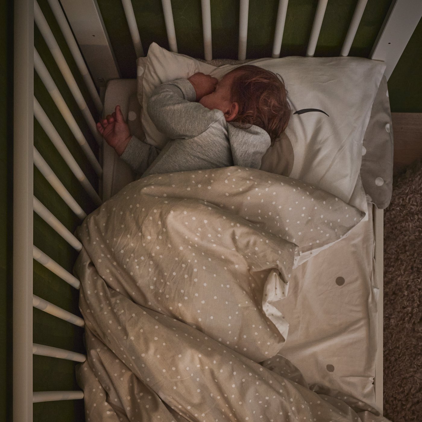 Ett litet barn sover fridfullt i en vit spjälsäng i trä, fotograferad uppifrån.