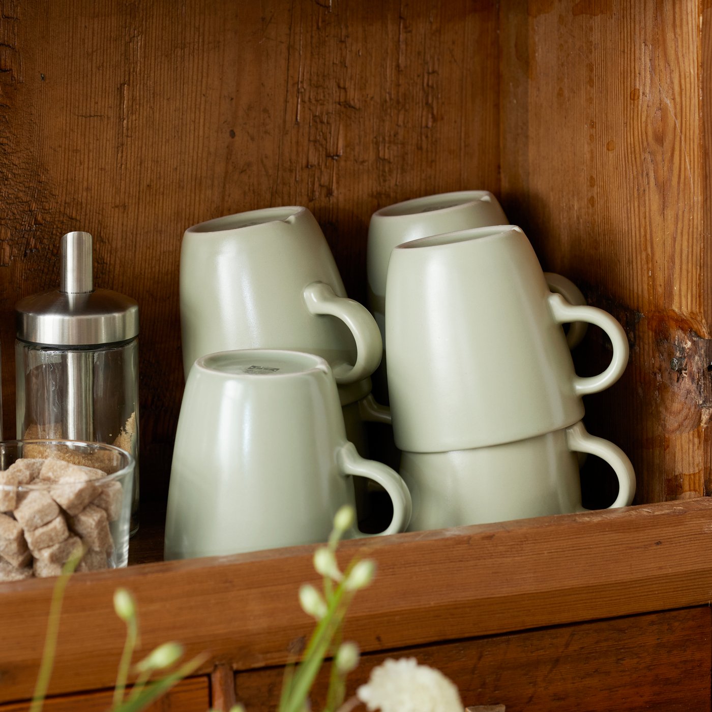A shelf of a wooden cabinet contains stacked FÄRGKLAR mugs, a glass with sugar cubes and a portion sugar shaker.