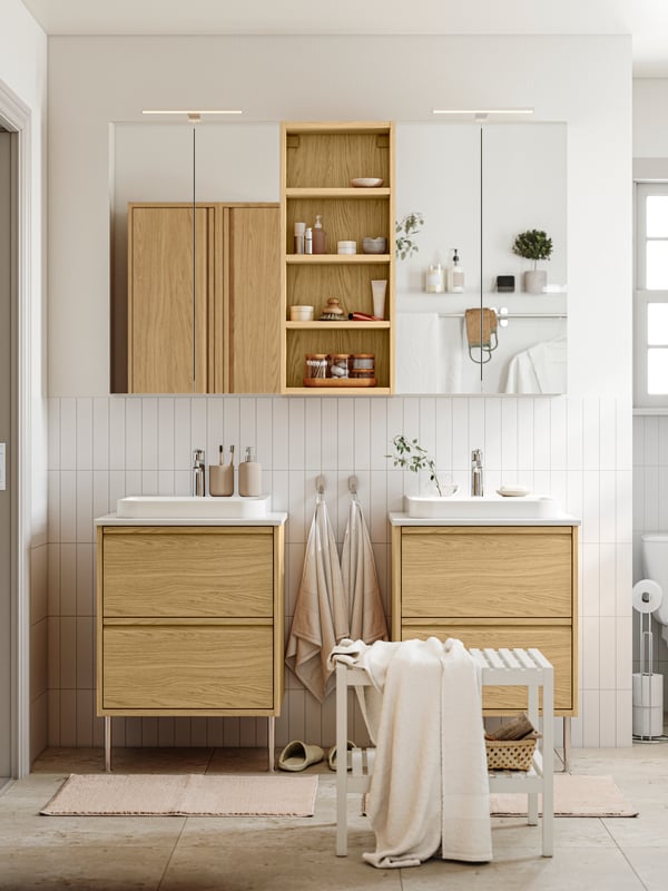 A white bathroom has ÄNGSJÖN bathroom furniture in oak effect, including two washstands with drawers and two mirror cabinets.