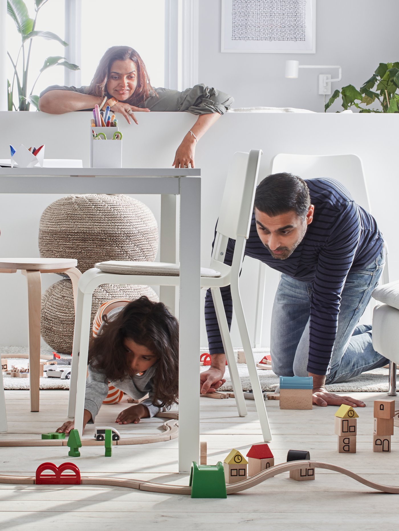 A dad down on his knees playing with his daughter under the dining room table while mum looks on from the sofa.
