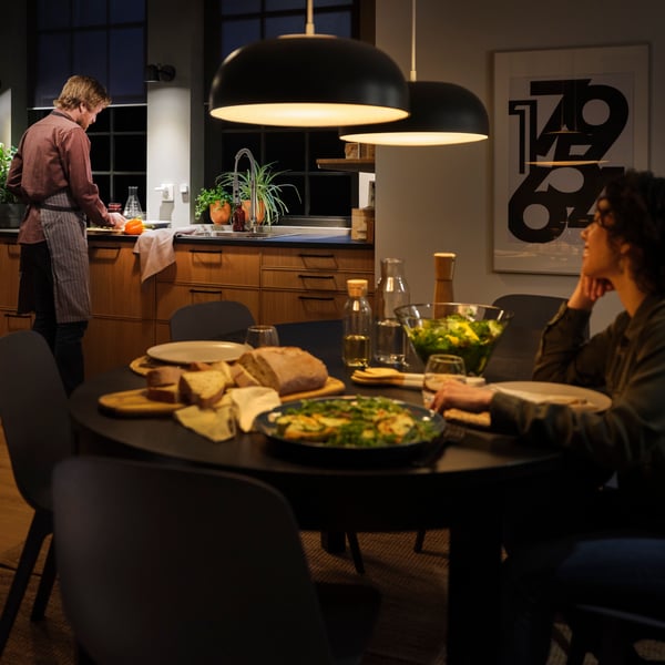A dinner table with dimmed lighting, a woman seated and a man cooking in the kitchen in the background.