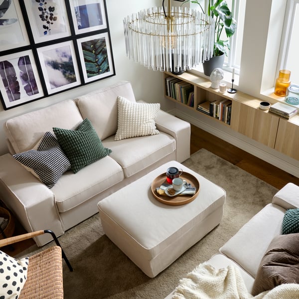 Ein Wohnzimmer mit zwei gegenüberstehenden KIVIK-Zweisitzsofas mit bunten Textilien, einem Pouf dazwischen und ordentlich angeordneten Wandbildern.