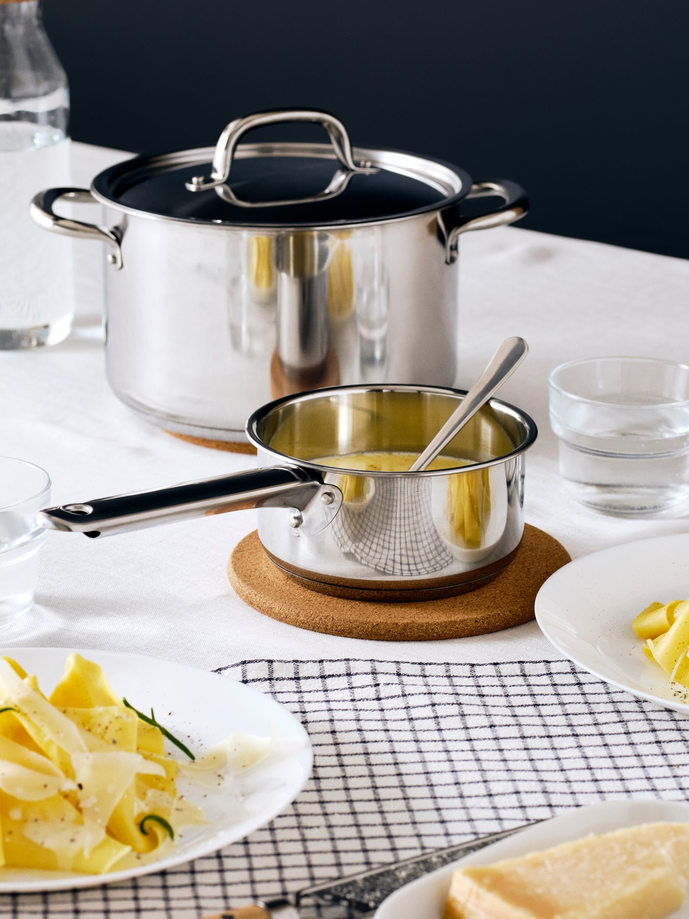 An IKEA365+ saucepan with lid in stainless steel on HEAT pot stand in cork and an IKEA365+ pot with lid in stainless steel.