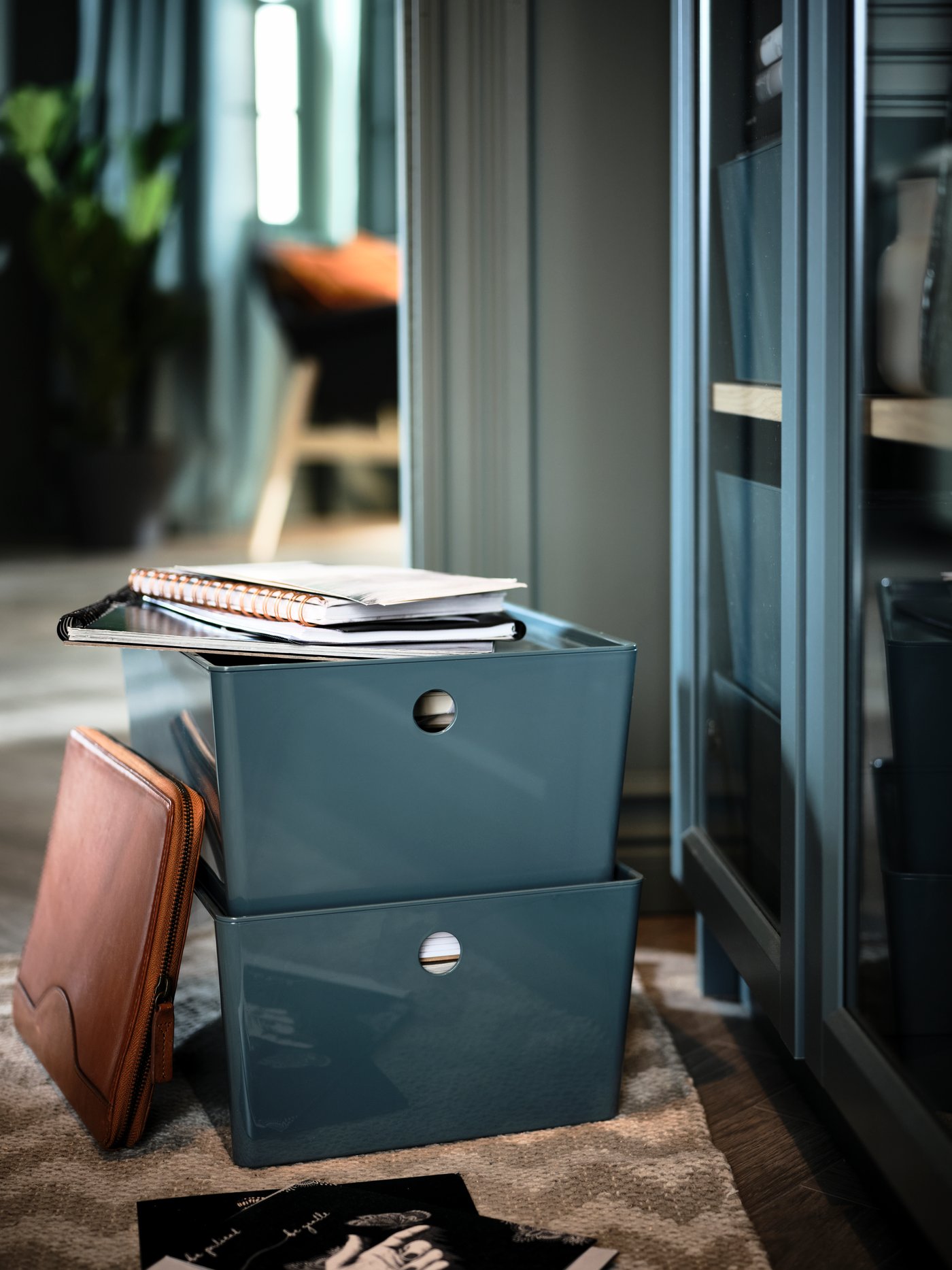 Two turquoise KUGGIS storage boxes holding notebooks and paper stand stacked on a rug beneath a BILLY glass-door bookcase.