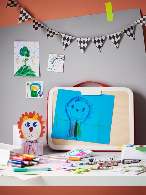 MÅLA Zeichnertasche mit Kinderbild, Buntstiften, Stiften und mehr auf einem Schreibtisch. An der Wand sind Zeichnungen und Wimpel zu sehen.