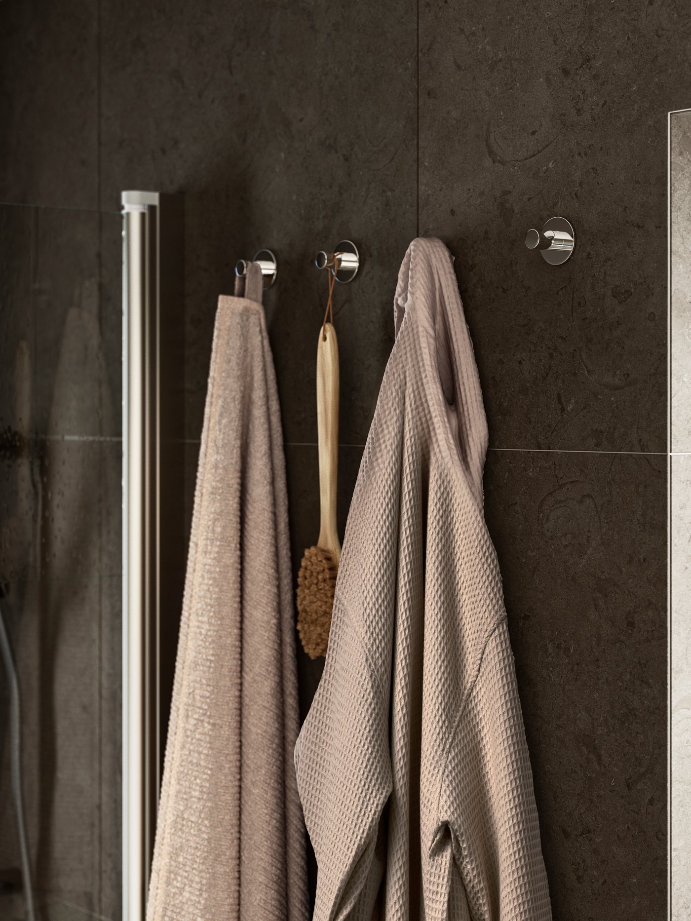 A beige, waffle-textured BJÄLVEN bathrobe hangs on a hook on the wall beside a beige VALLASÅN towel and a wooden body brush.