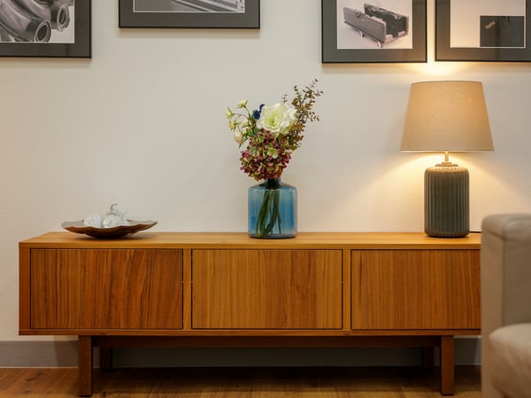 木製サイドボードの上に花瓶とテーブルランプを置いたリビングの一角。壁にアートを飾ったあたたかみのあるインテリア。