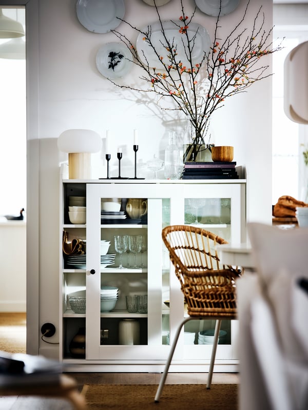 A softly sunlit, white dining area flanked by a white HAUGA glass-door cabinet with tableware in it and decoration on it.