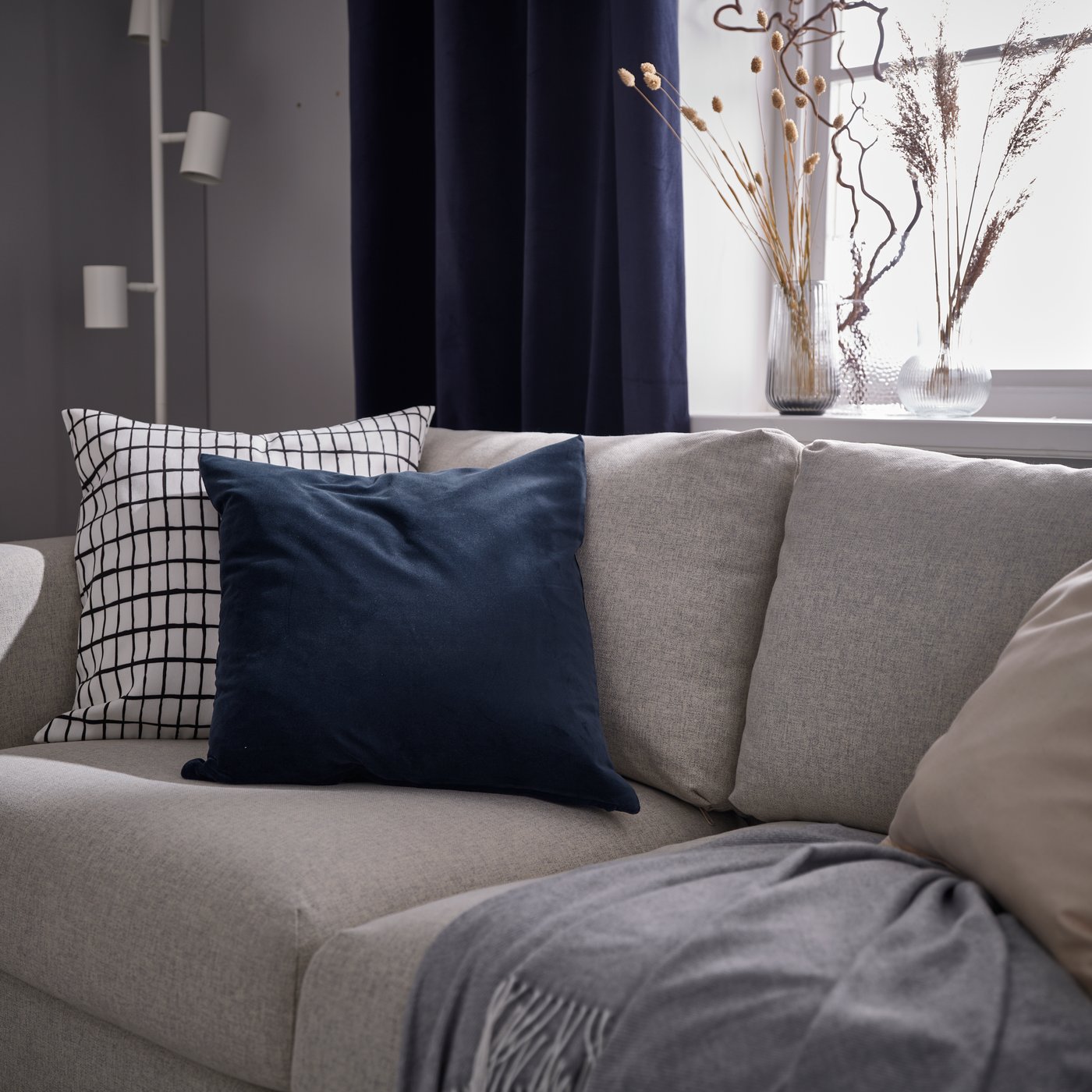 Auf einem beigen VIMLE Sofa liegen ein blaues SANELA Kissen aus Samt, ein weiß/schwarz kariertes MÄSTERROT Kissen und ein graues HOLMVI Plaid aus recyceltem Polyester.