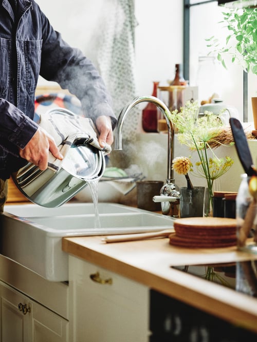 O persoană ține un vas IKEA 365+ cu capac peste un bol de chiuvetă HAVSEN cu un robinet de mixer INSJÖN. Oala are conținut de abur.