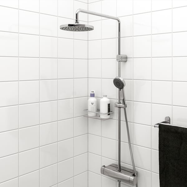 A modern shower head attached to a sleek, vertical bar mounted on a wall with neutral tones. A green leaf from an out-of-focus plant in the foreground adds a touch of nature to the scene. Below the shower head, on an integrated shelf, rests a natural loofah and green toiletries, suggesting a bathroom setting focused on relaxation and cleanliness. The background wall is subtly textured, complementing the simple yet elegant design of the shower fixture.