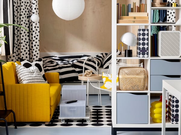 A FRIDHULT sofa-bed stands in a living room area near a white KALLAX shelving unit and a bed with striped bed linen.