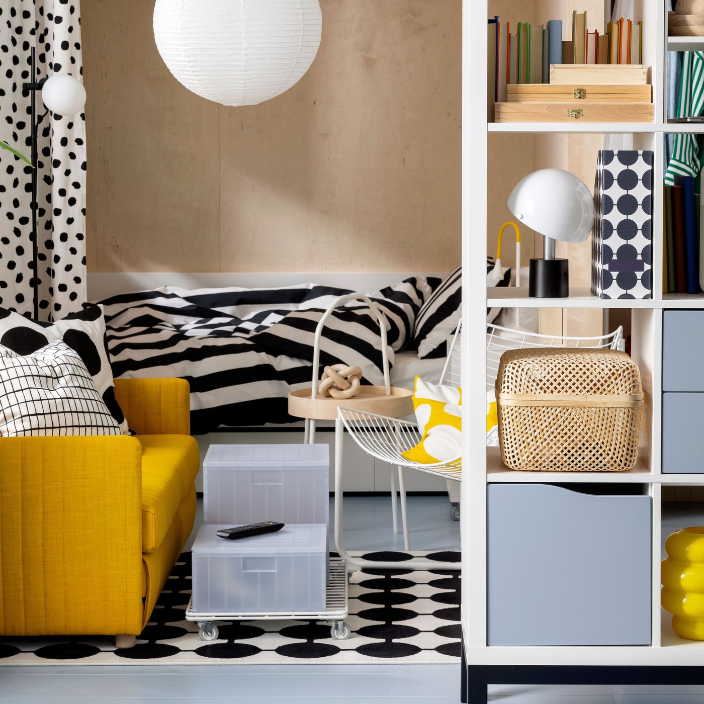 A FRIDHULT sofa-bed stands in a living room area near a white KALLAX shelving unit and a bed with striped bed linen.