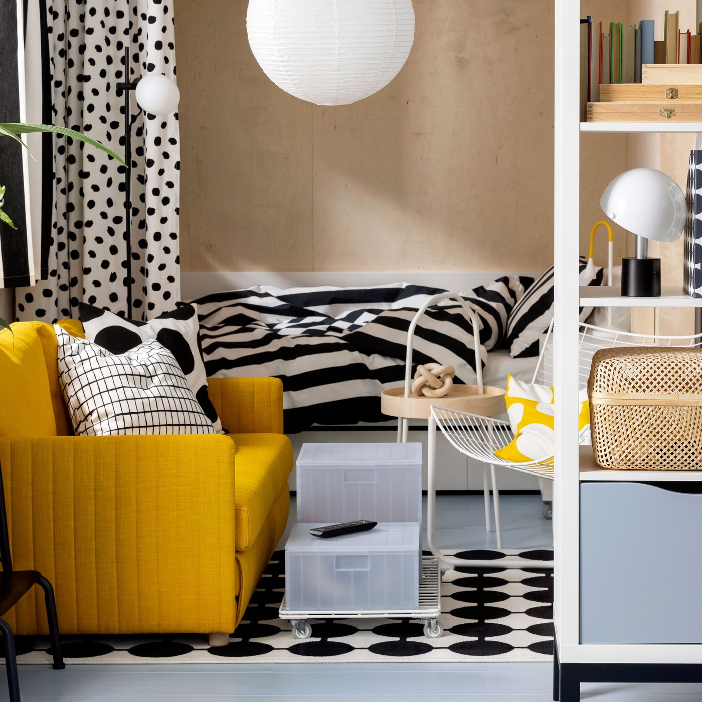 IKEA FRIDHULT yellow sofa-bed stands in a living room area near a white KALLAX shelving unit and a bed with striped bed linen.