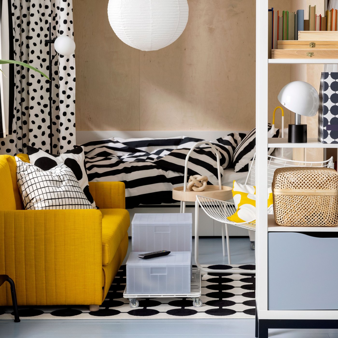 En FRIDHULT bäddsoffa står i ett vardagsrumsområde nära en vit KALLAX hyllningsenhet och en säng med randigt sänglinne.