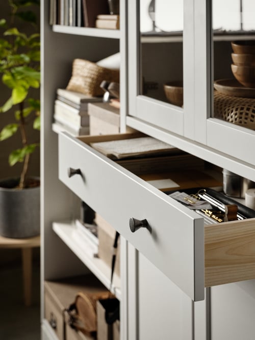 A white high cabinet with glass doors and 1 drawer, a white bookcase, books, storage boxes with lids.