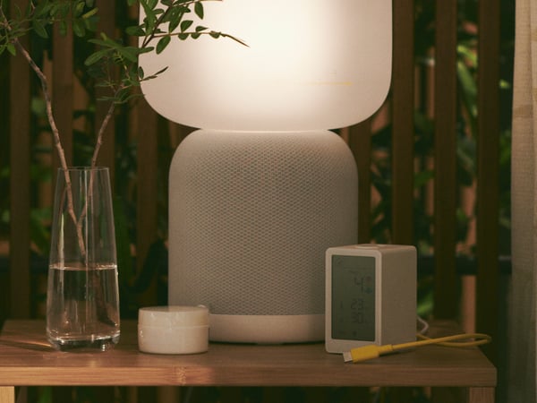 A SYMFONISK table lamp with WiFi speaker in white/grey placed on a bedside table.