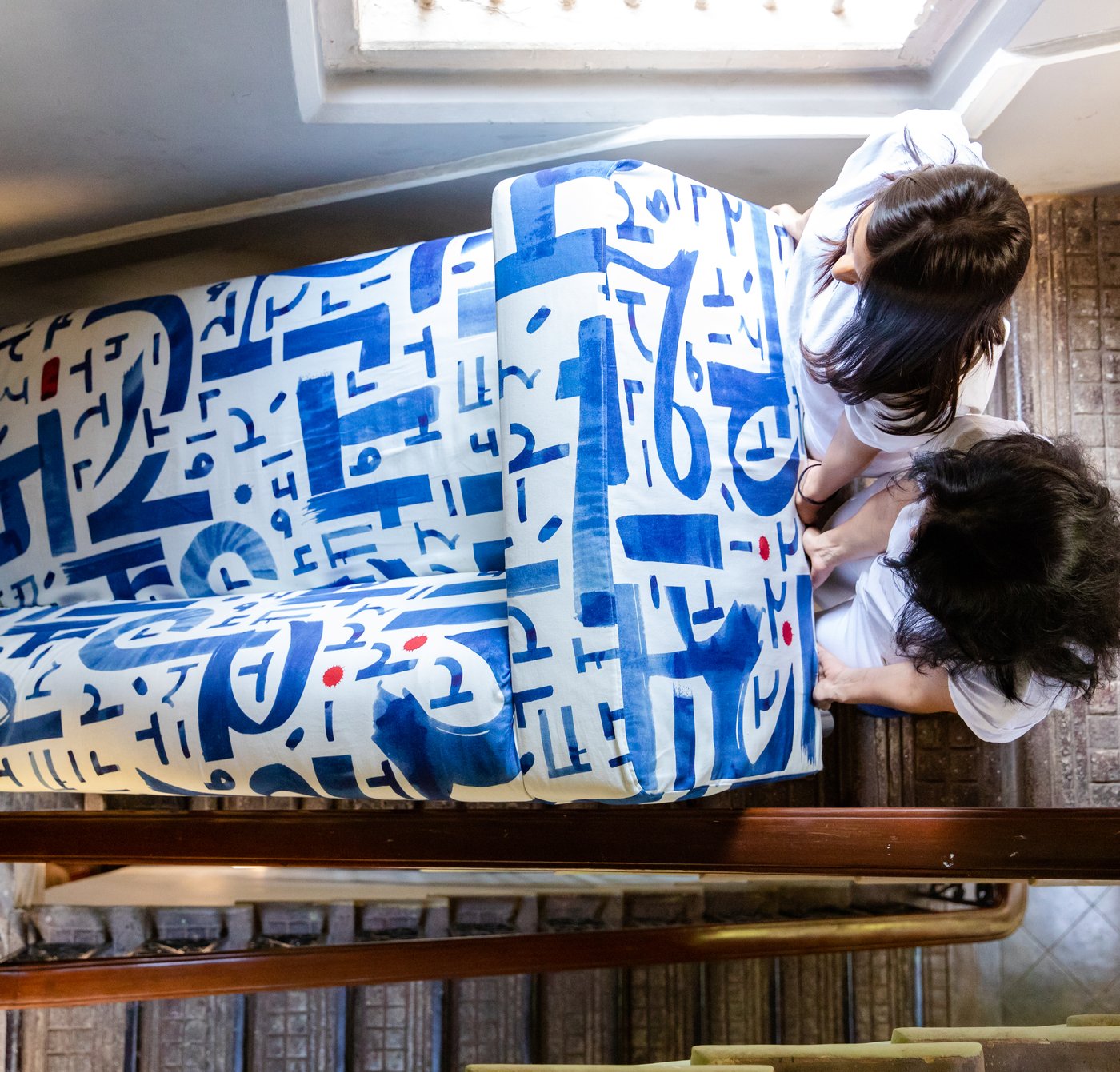 To kvinner bærer en sofa med blått og hvitt tekstiltrekk i ei trapp ved siden av et vindu.