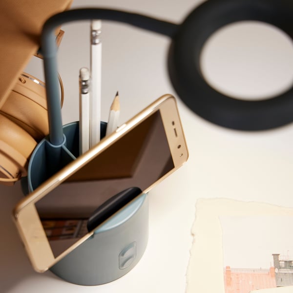 A smart phone sits horizontally in the phone holder of a dimmable, turquoise BRUNBÅGE LED work lamp with storage.