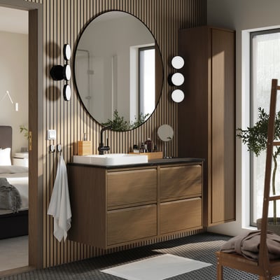 Modern bathroom: round mirror, oak vanity, marble basin, wall-mounted, plant, towels, basket, patterned floor, bed beyond.