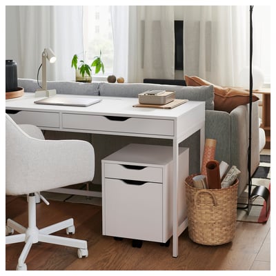 Modern home office setup with white ALEX desk and chair, featuring a laptop, basket with supplies, and a cozy sofa with a cushion and blanket.