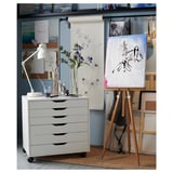 White storage unit with five drawers, wooden legs, used in an artists studio next to an easel and paintings.