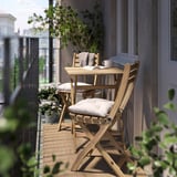 Small balcony with light wooden table and chairs, cushioned seats, potted plants, and coffee cup.