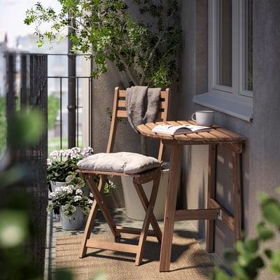 Small balcony setup with wooden chair & table, grey cushion & blanket, white cup, book & potted plant.