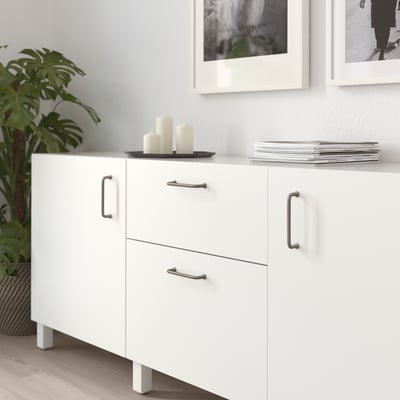 White BAGGANÄS cabinet with metal handles on wood floor, frame, plant.