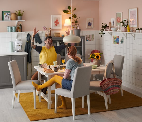 Two people dining at modern table with yellow and gray BERGMUND chairs, surrounded by plants, paintings, and a unique pendant lamp.