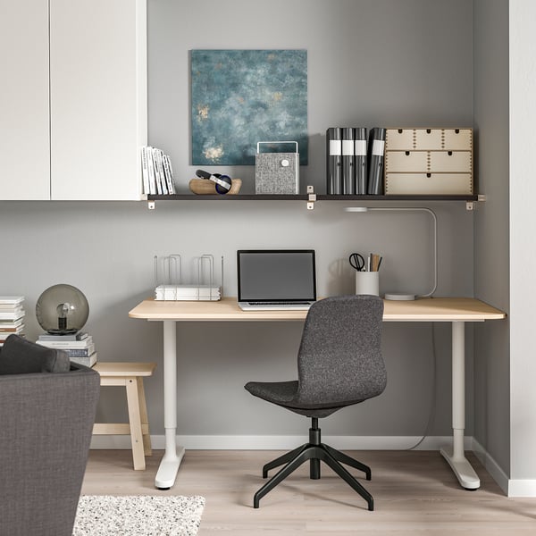 Home office setup: white desk, gray chair, laptop, wall shelf with books, art, light.