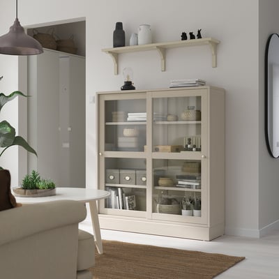 BERGSHULT beige shelf with glass doors, holding items, under wall shelf, near sofa and plant table.