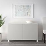 A white sideboard with two doors and a top surface holding a bowl. A framed art piece hangs above.