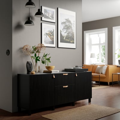 Dark wood BESTÅ sideboard holds & displays items. Three framed photos hang above. Orange sofa in living area beside window.
