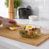 Person preparing a salad in a glass container on a wooden board. The container is suitable for stacking and storing food.