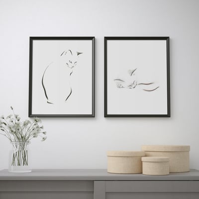 Two black-framed cat artwork pieces hang above a table with a vase of flowers and woven baskets.