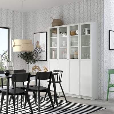 Modern dining area with white BILLY bookcase, black chairs, striped rug, geometric wallpaper, and potted plant.